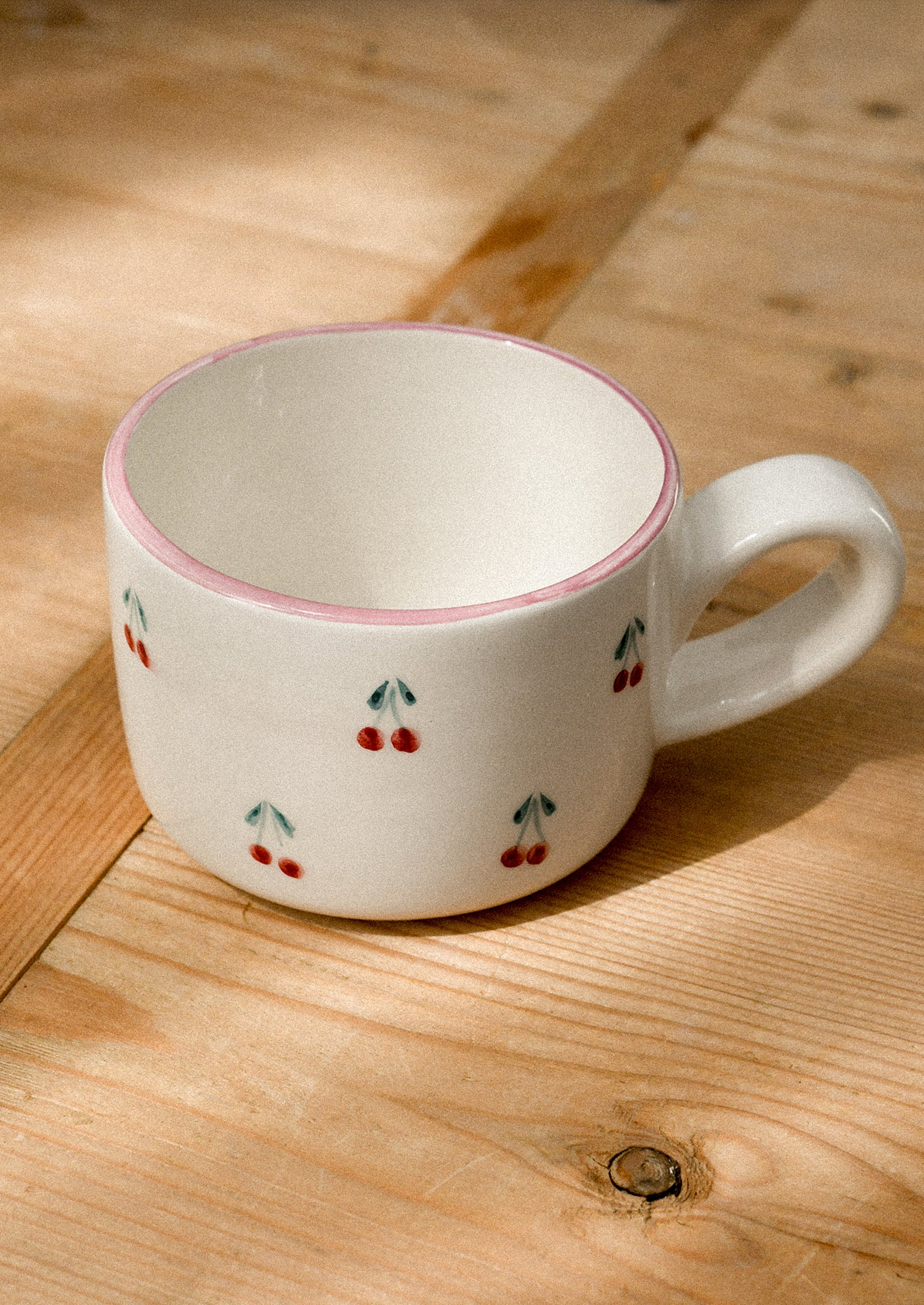 A cream ceramic mug with hand painted cherry motif and pink top rim.