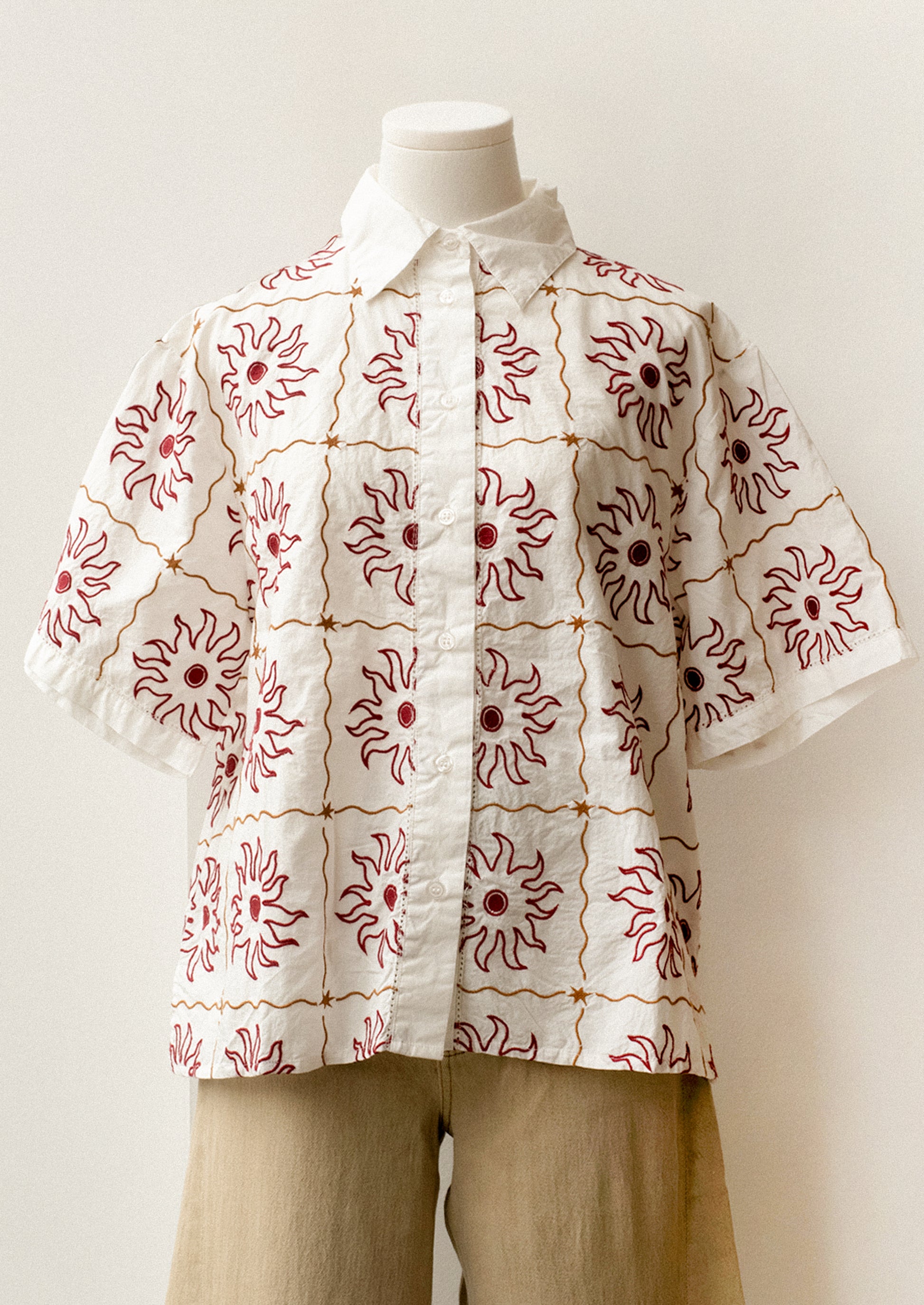 A short sleeve, collared shirt in white with red and brown sun embroidery.