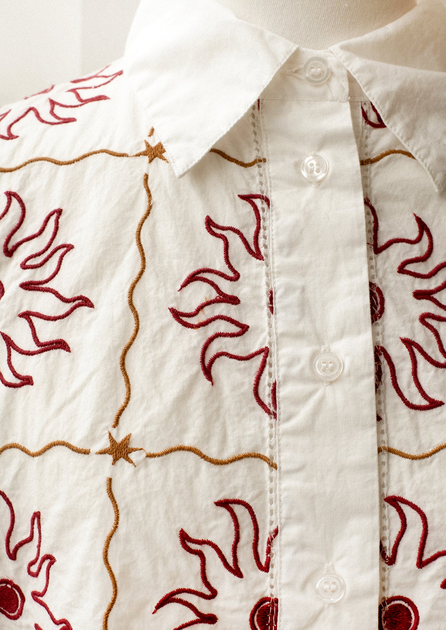 A short sleeve, collared shirt in white with red and brown sun embroidery.