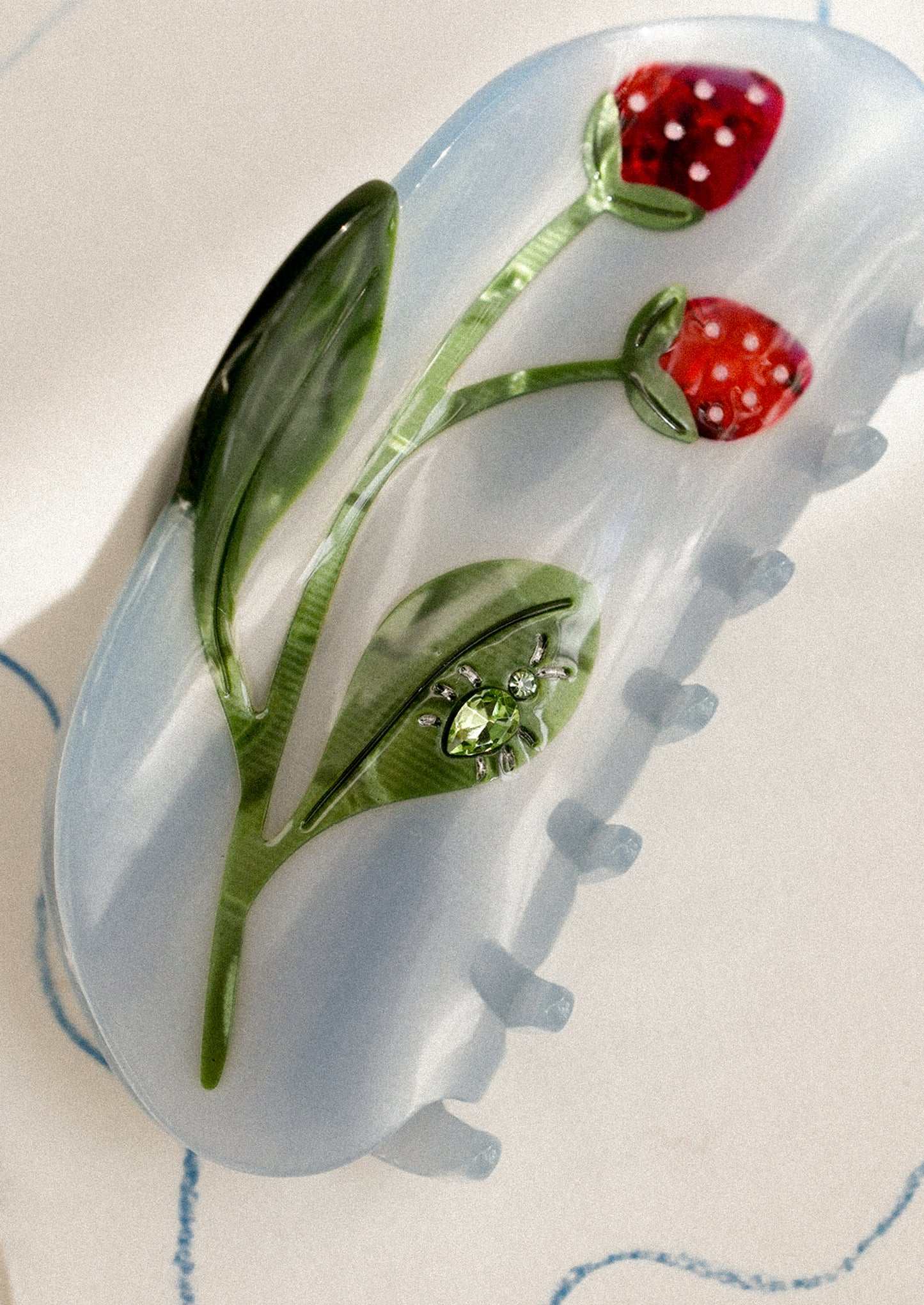 A blue hair claw with strawberry motif and jeweled bug on leaf.