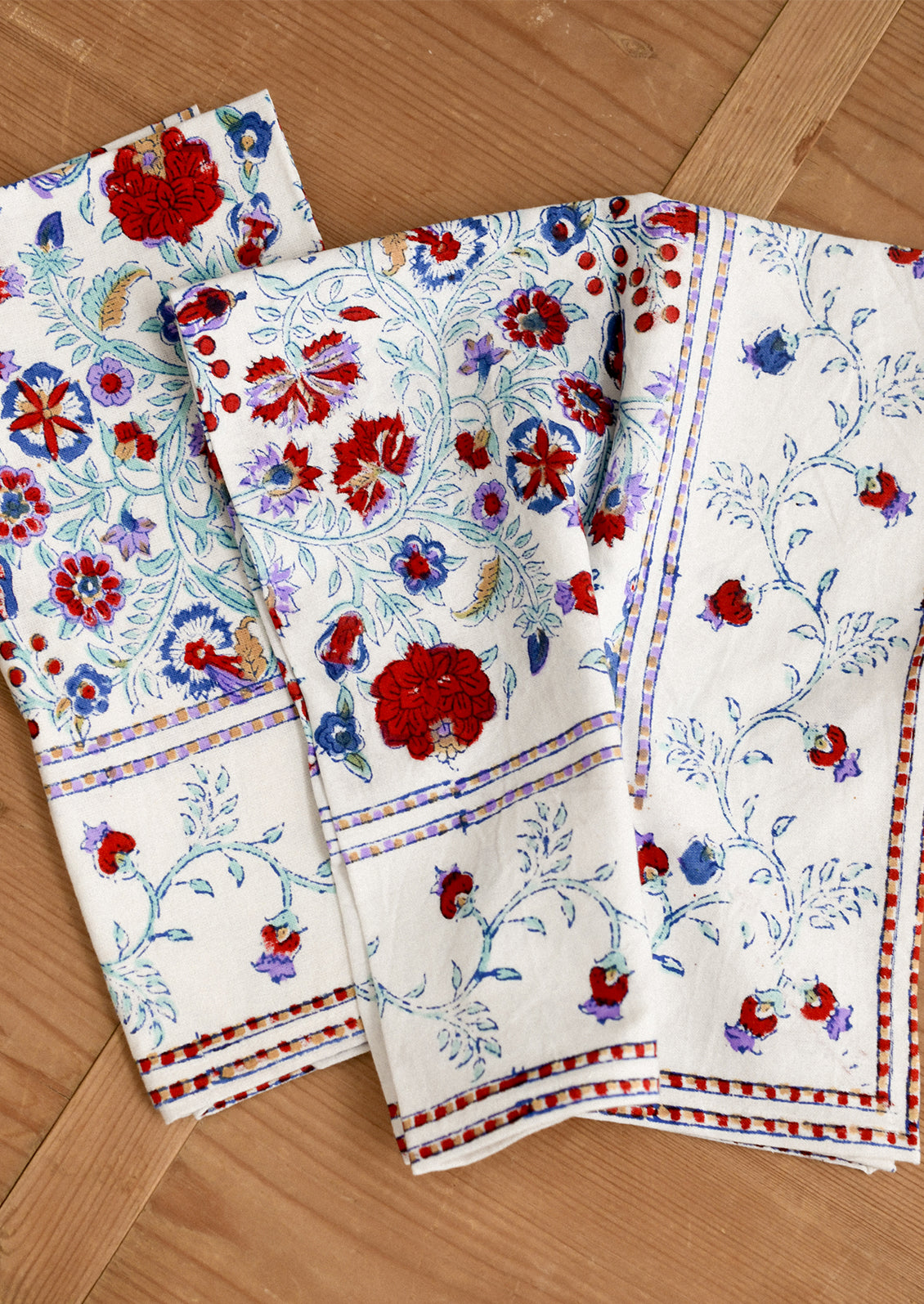 Set of floral-patterned napkins in white with red, lavender and aqua color scheme on a wooden surface.