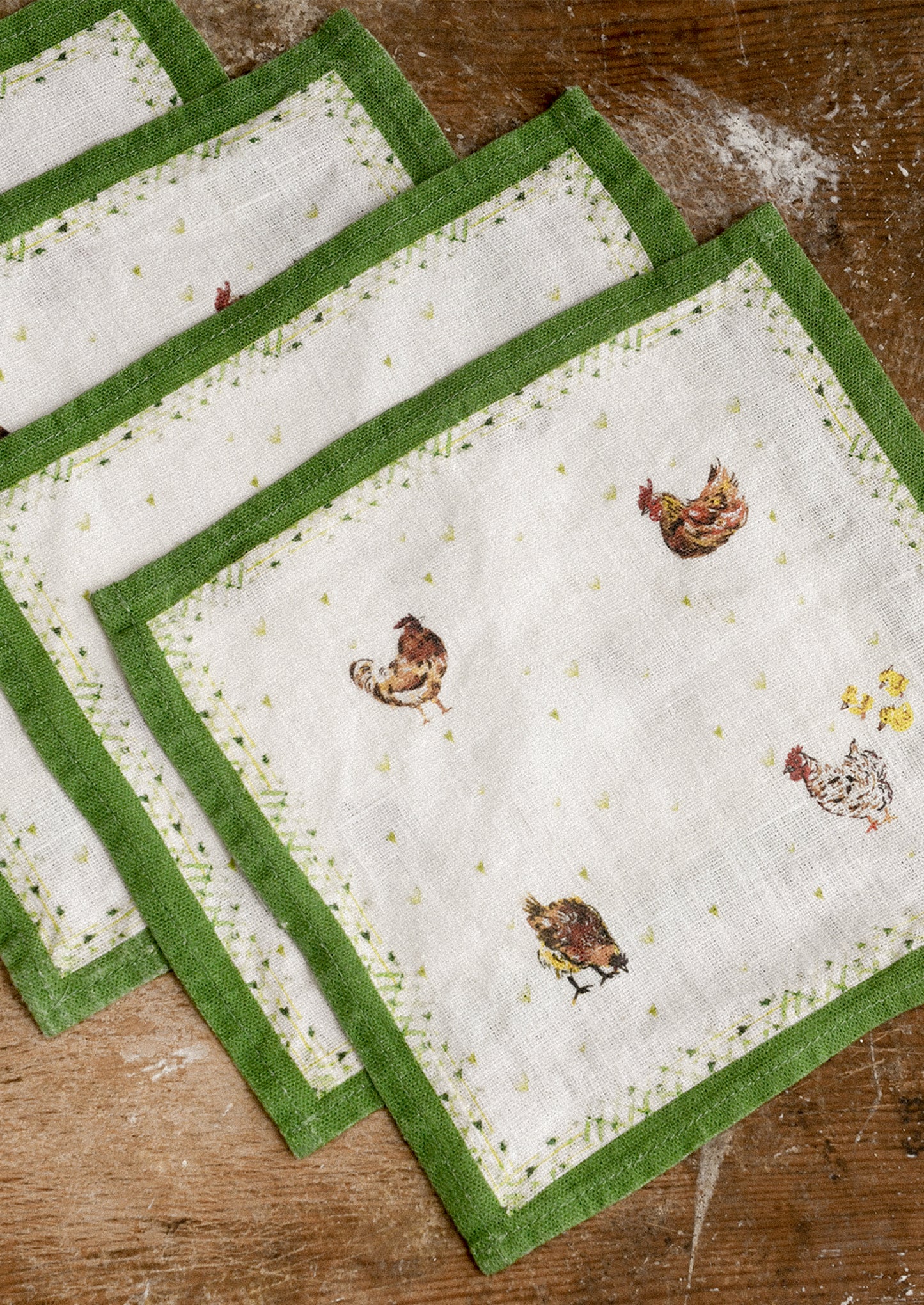Set of four cocktail napkins with green borders and chicken designs on a wooden surface