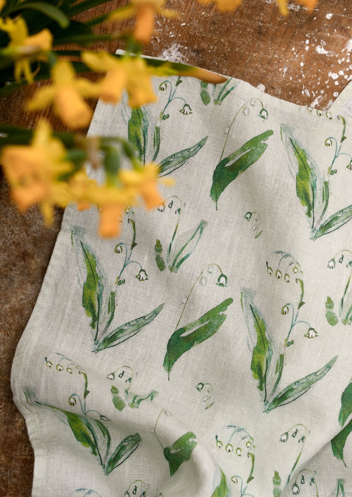 Kitchen tea towel with green lily of the valley pattern on a wooden surface with yellow flowers.