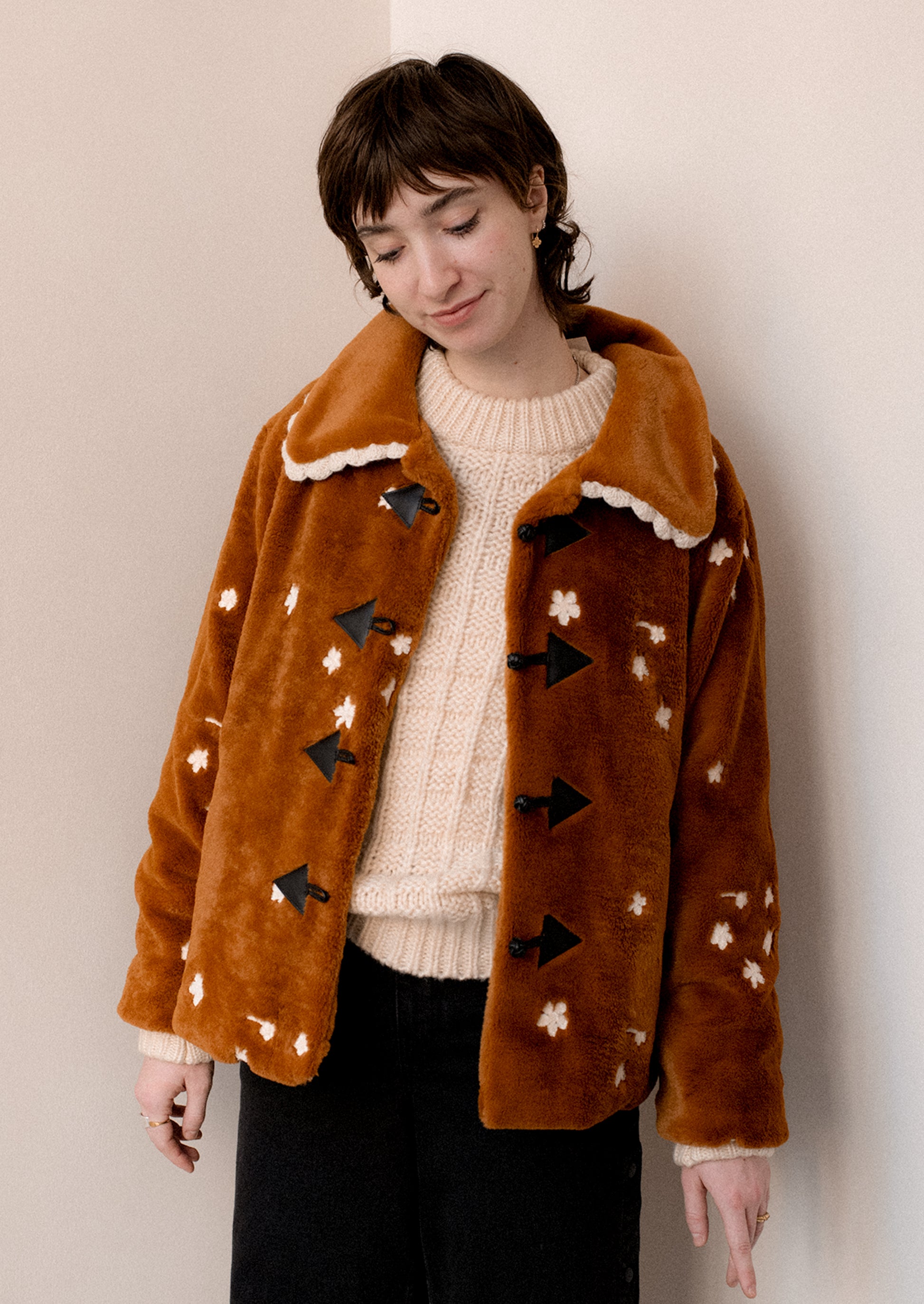 A woman wearing a caramel colored faux fur jacket with white floral embroidery and black toggle button front.