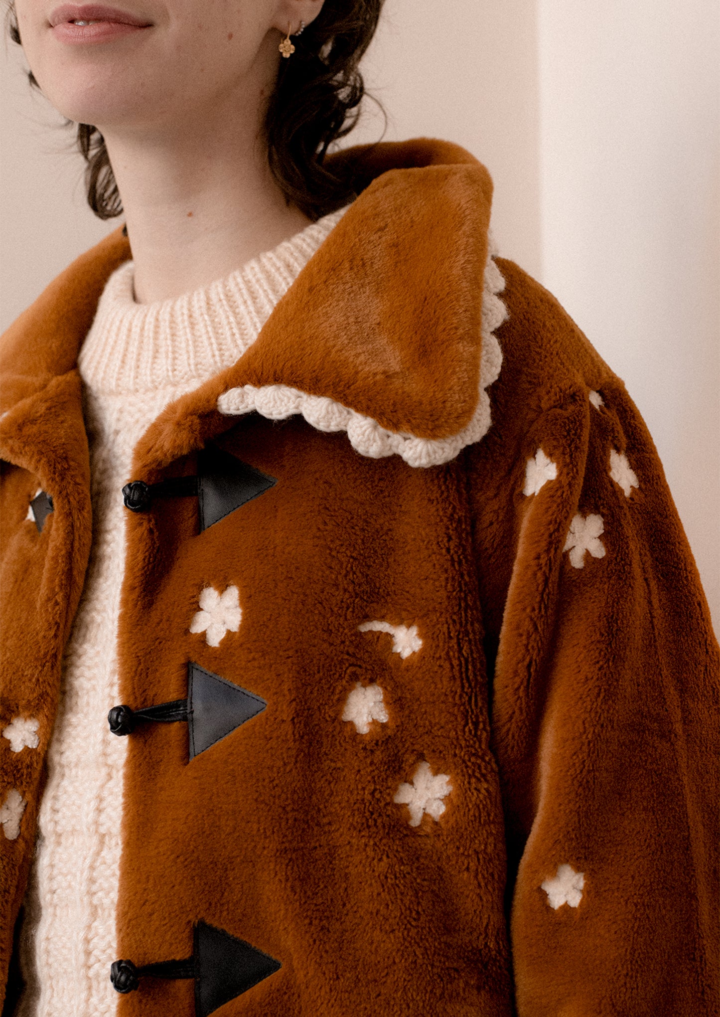 A woman wearing a caramel colored faux fur jacket with white floral embroidery and black toggle button front.