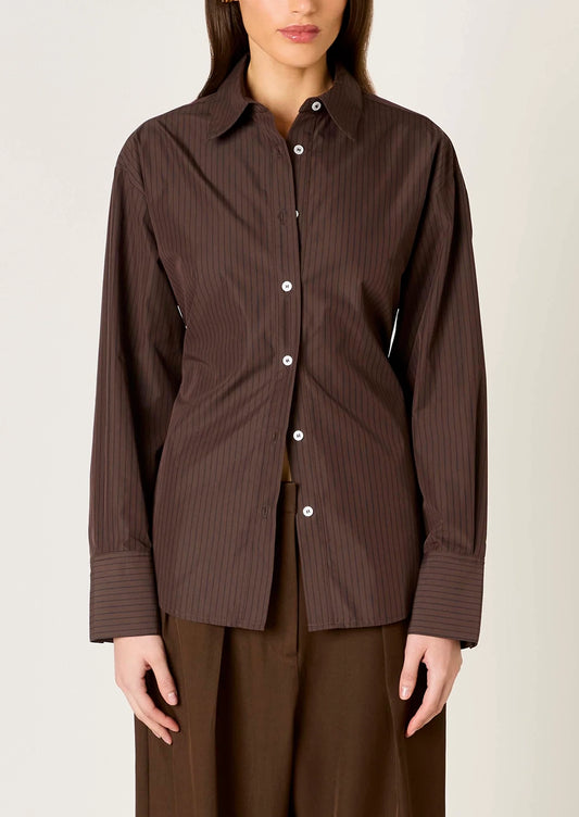A woman wearing a dark brown long-sleeve button-up shirt with thin vertical pinstripes and white buttons, paired with matching brown trousers, against a plain light beige background.