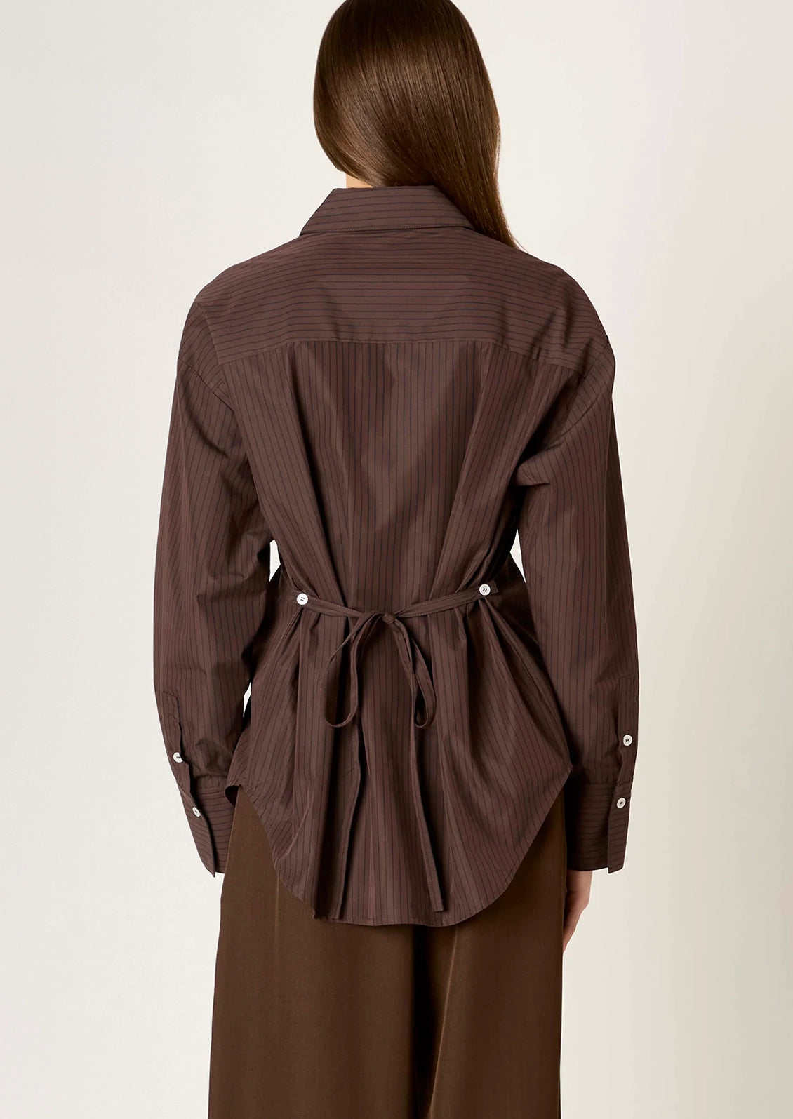 A woman viewed from the back wearing a dark brown pinstriped long-sleeve shirt with a tie-waist detail and brown trousers, set against a plain off-white studio background.