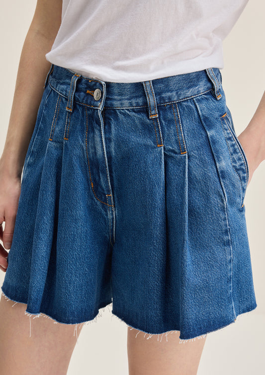A person wearing a pair of pleated denim shorts in true medium blue wash.