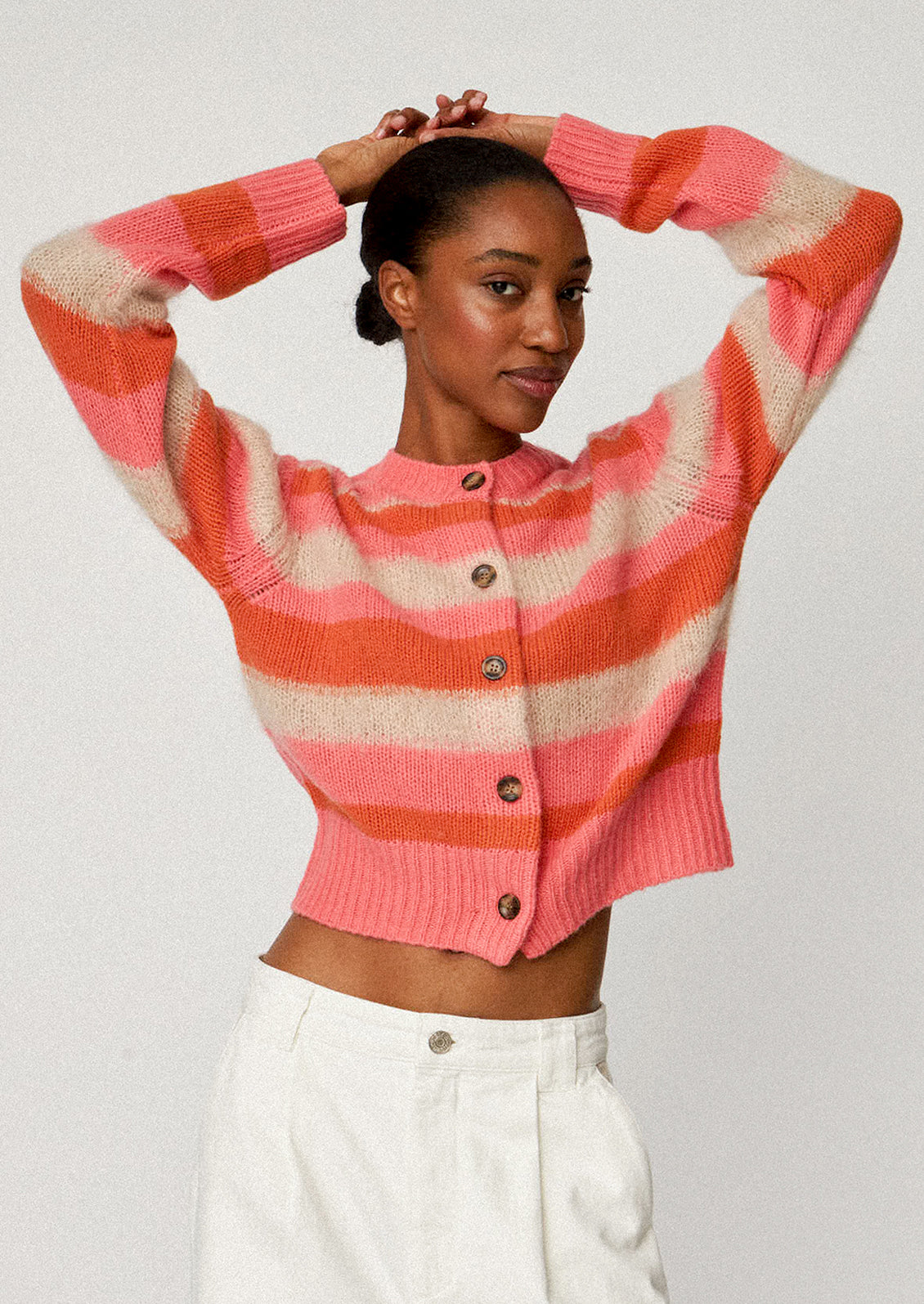 A woman wearing a button front cardigan sweater in pink, orange and ivory stripes.