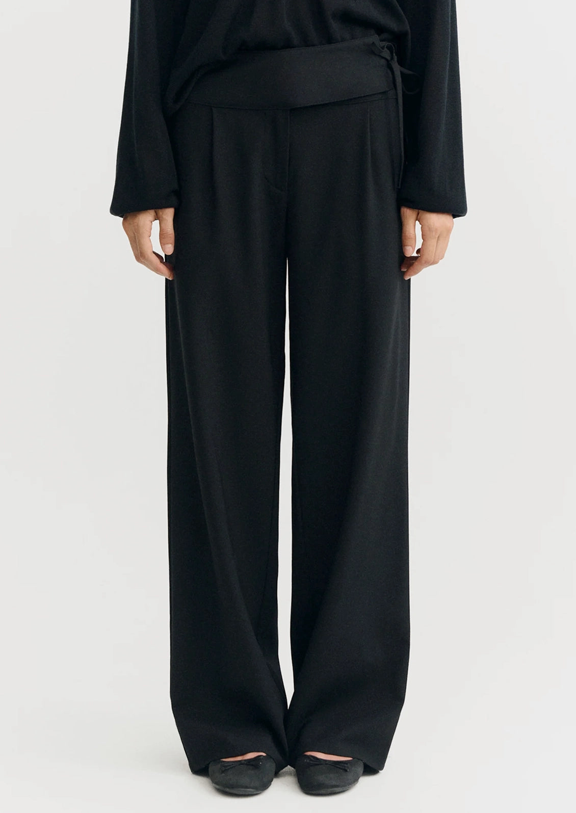 A model wearing black wide-leg trousers with a matching black top, shown from the waist down against a plain light gray studio background.