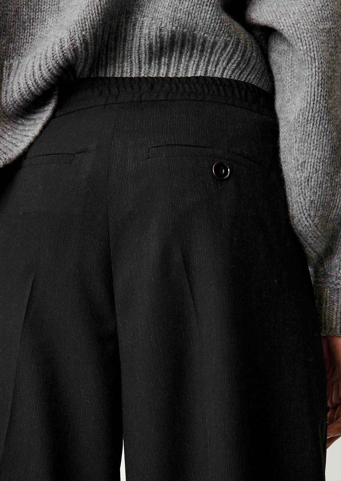 Close-up of the back waist of a person wearing black pleated trousers and a gray ribbed knit sweater.