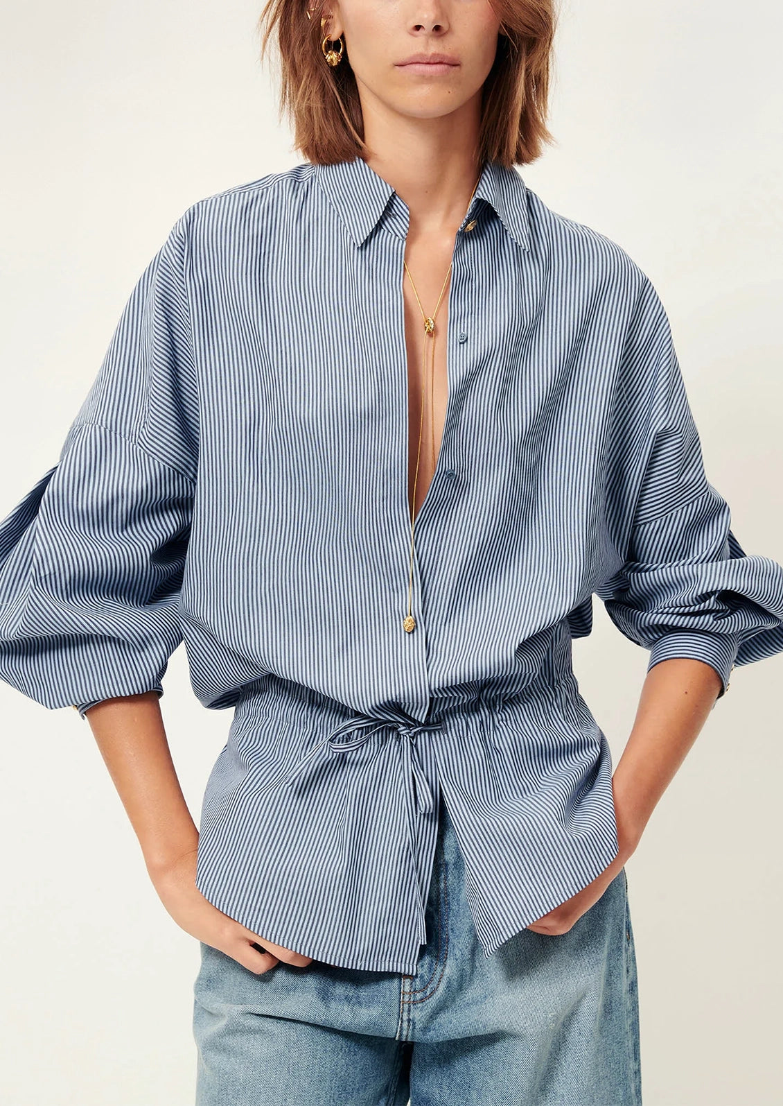 Woman wearing a blue striped shirt with a tie waist and light blue pants on a white background