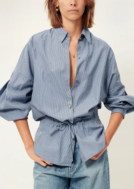 Woman wearing a blue striped shirt with a tie waist and light blue pants on a white background