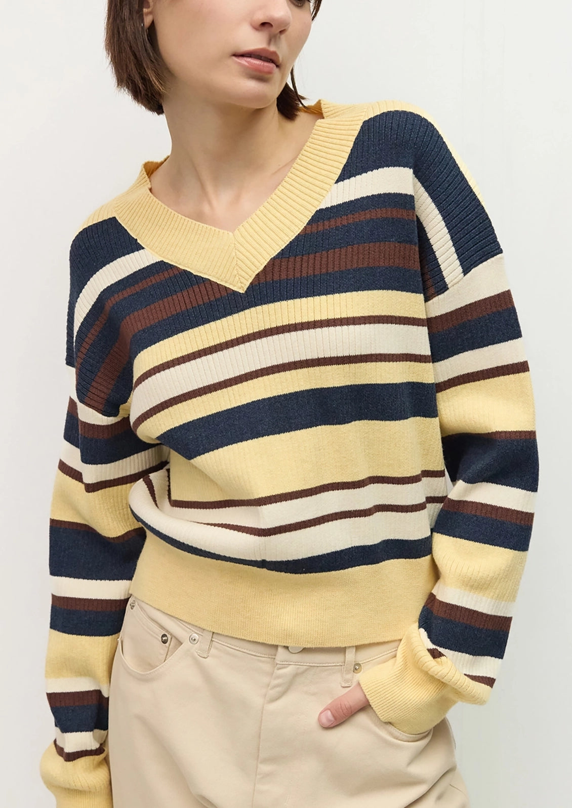 A woman wearing a long-sleeve V-neck knit sweater with horizontal stripes in pale yellow, navy blue, cream, and brown.