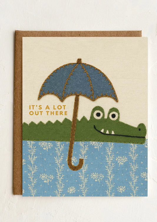 Card with an illustration of a crocodile holding an umbrella and text 'It's a lot out there' on a white background.