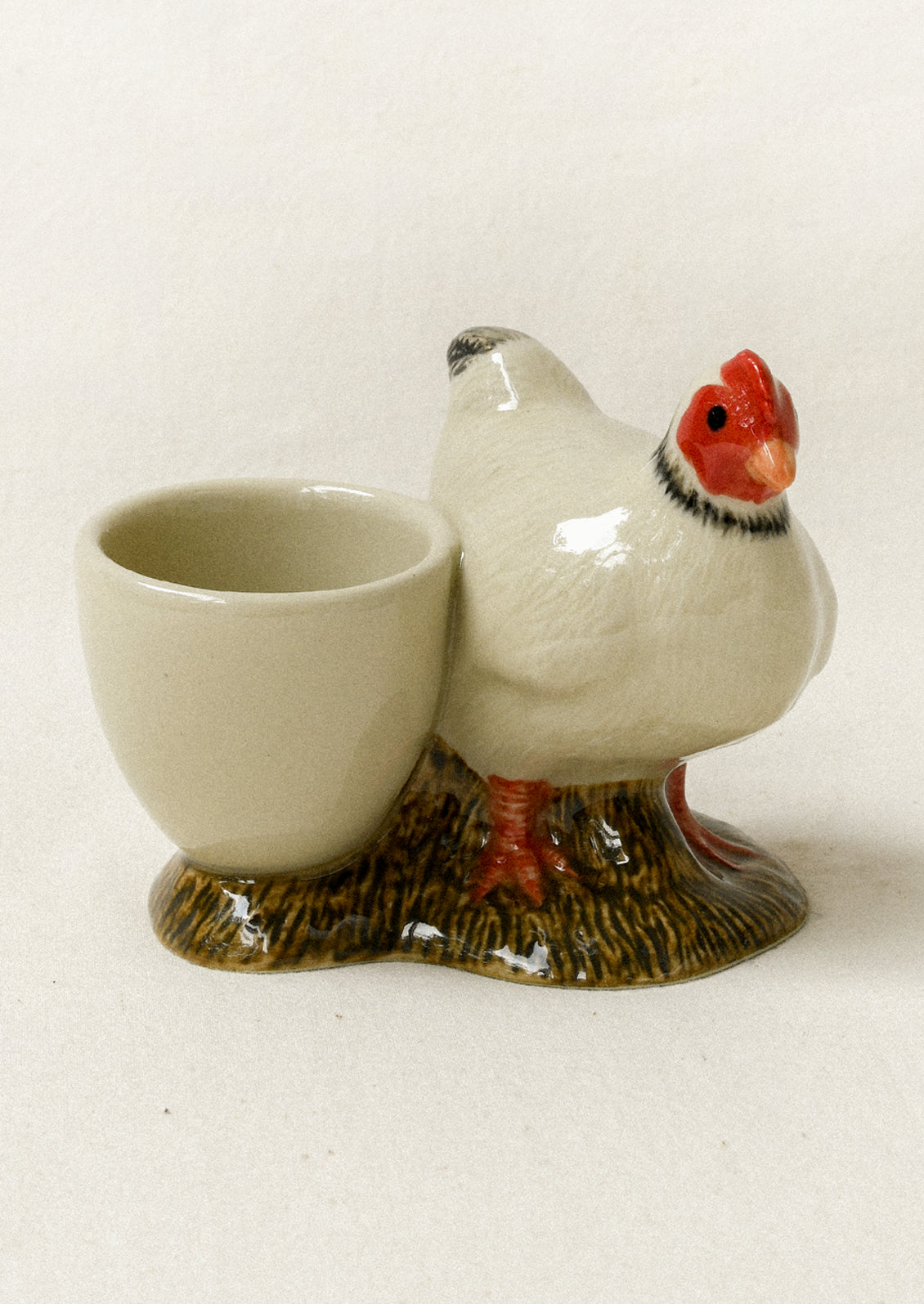 A ceramic egg cup holder shaped like a white hen standing on a brown textured ground, with a small off-white cup attached to its side.
