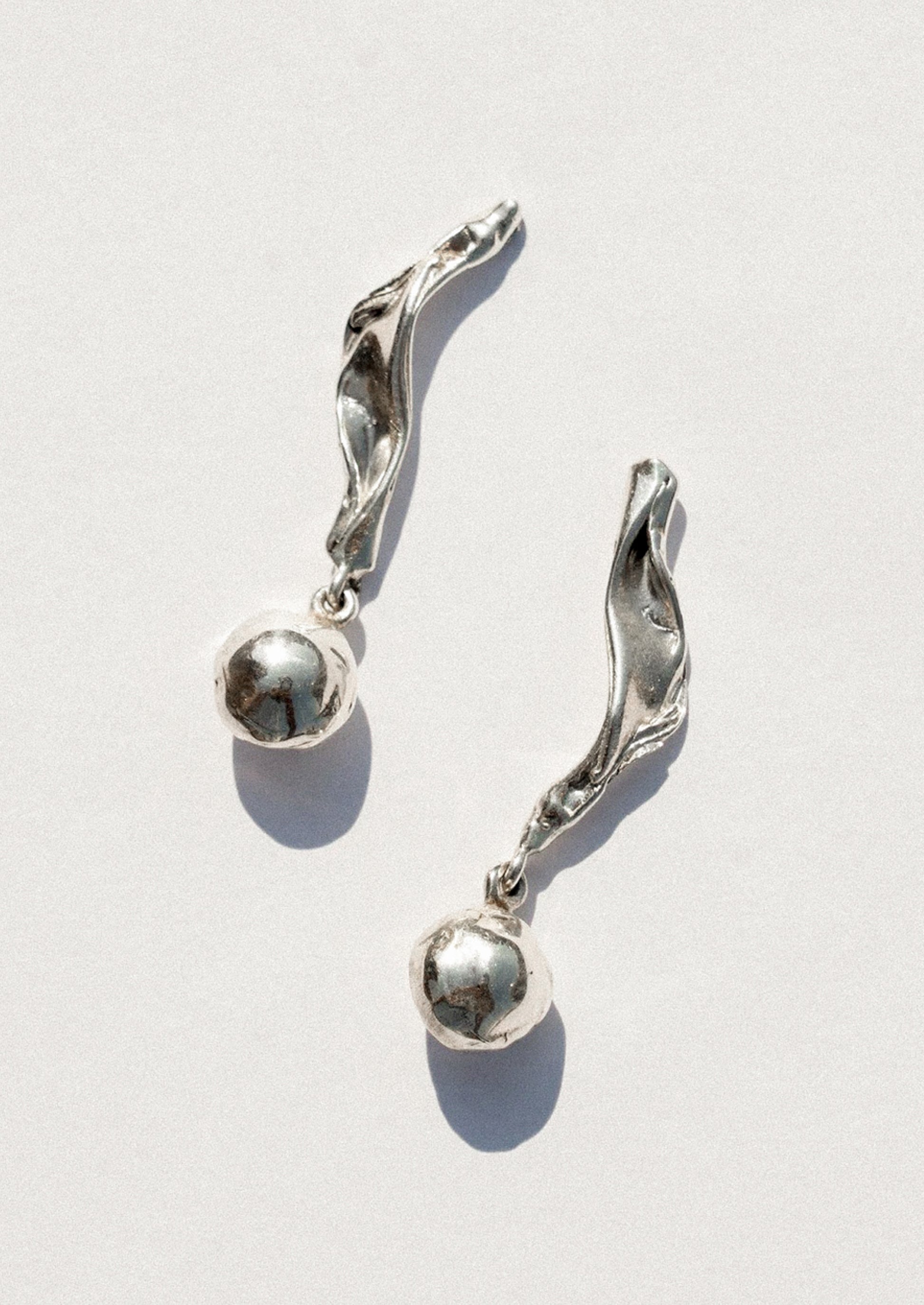 A pair of silver dangle earrings with textured, wave-like curved tops and smooth spherical beads hanging below, displayed on a clean white background with soft shadows.