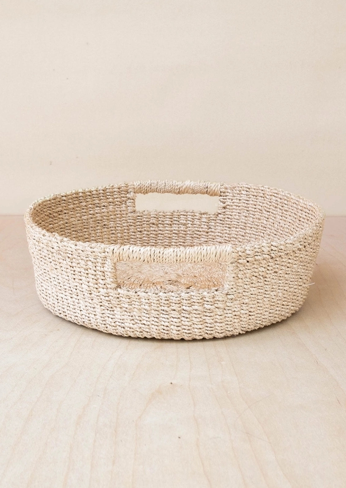 A round woven natural fiber basket with integrated handles, shown on a light wood surface against a neutral beige background.