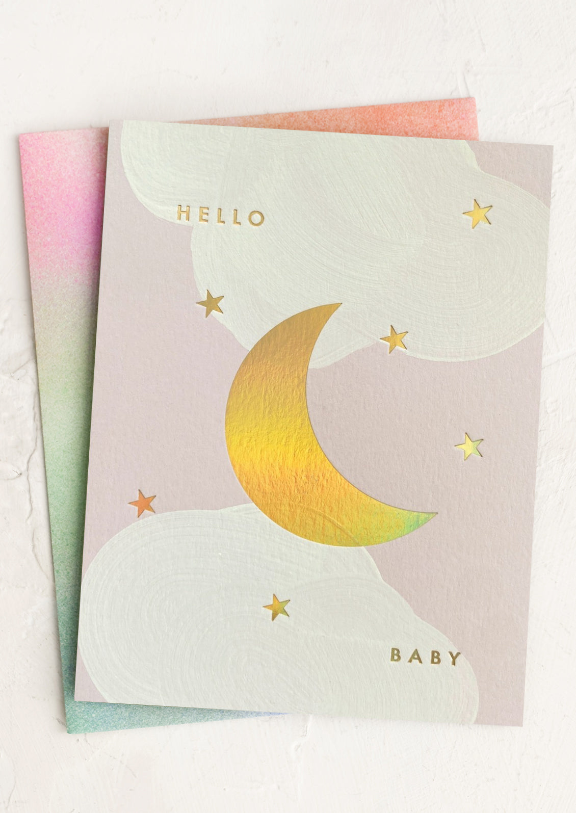 A greeting card with a gold holographic moon and stars design, with the text 'Hello Baby' on the front. The card is fanned out to show the holographic detail and includes a printed envelope.