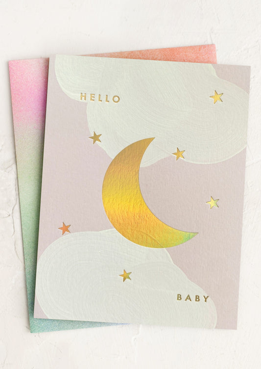 A greeting card with a gold holographic moon and stars design, with the text 'Hello Baby' on the front. The card is fanned out to show the holographic detail and includes a printed envelope.