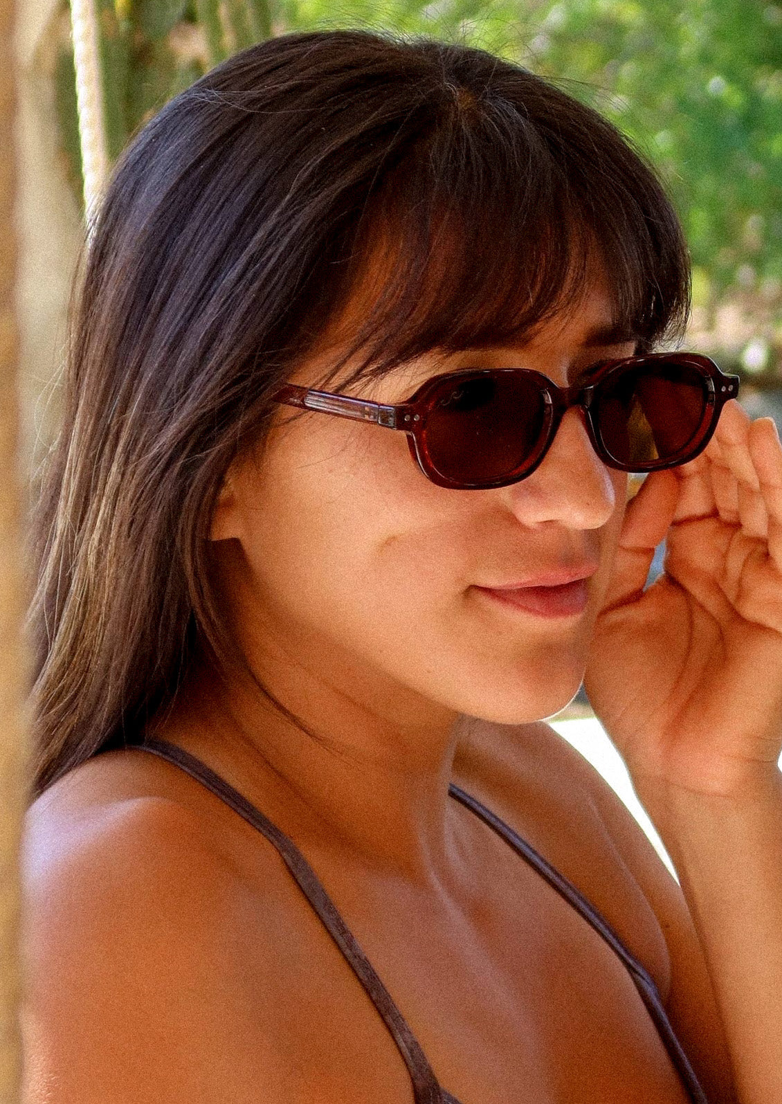 A woman wearing a pair of oval-rectangle sunglasses with a translucent cherry red frame and warm medium-brown tinted lenses.