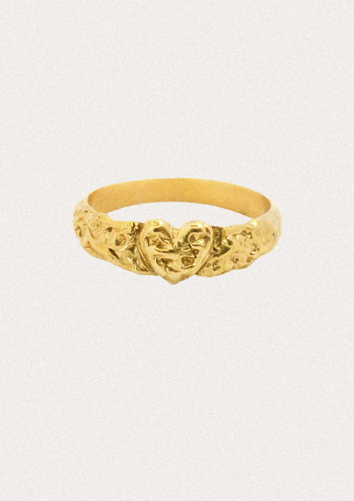 A textured gold ring featuring a central heart motif flanked by floral details, set against a plain light gray background.