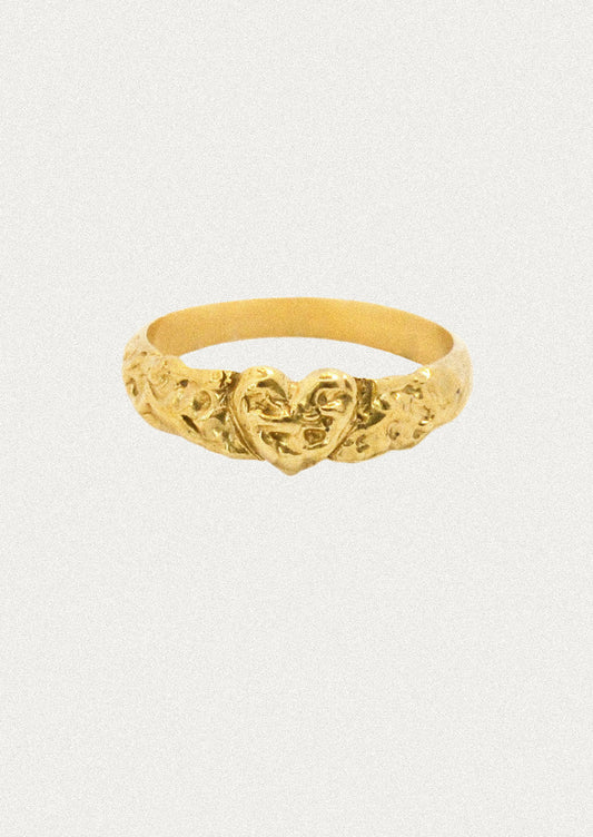 A textured gold ring featuring a central heart motif flanked by floral details, set against a plain light gray background.