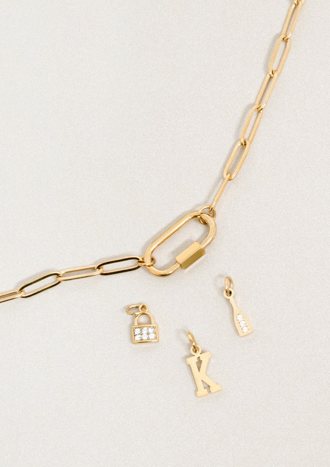 A gold chain necklace with a small functional carabiner and 3 small charms on a grey background.