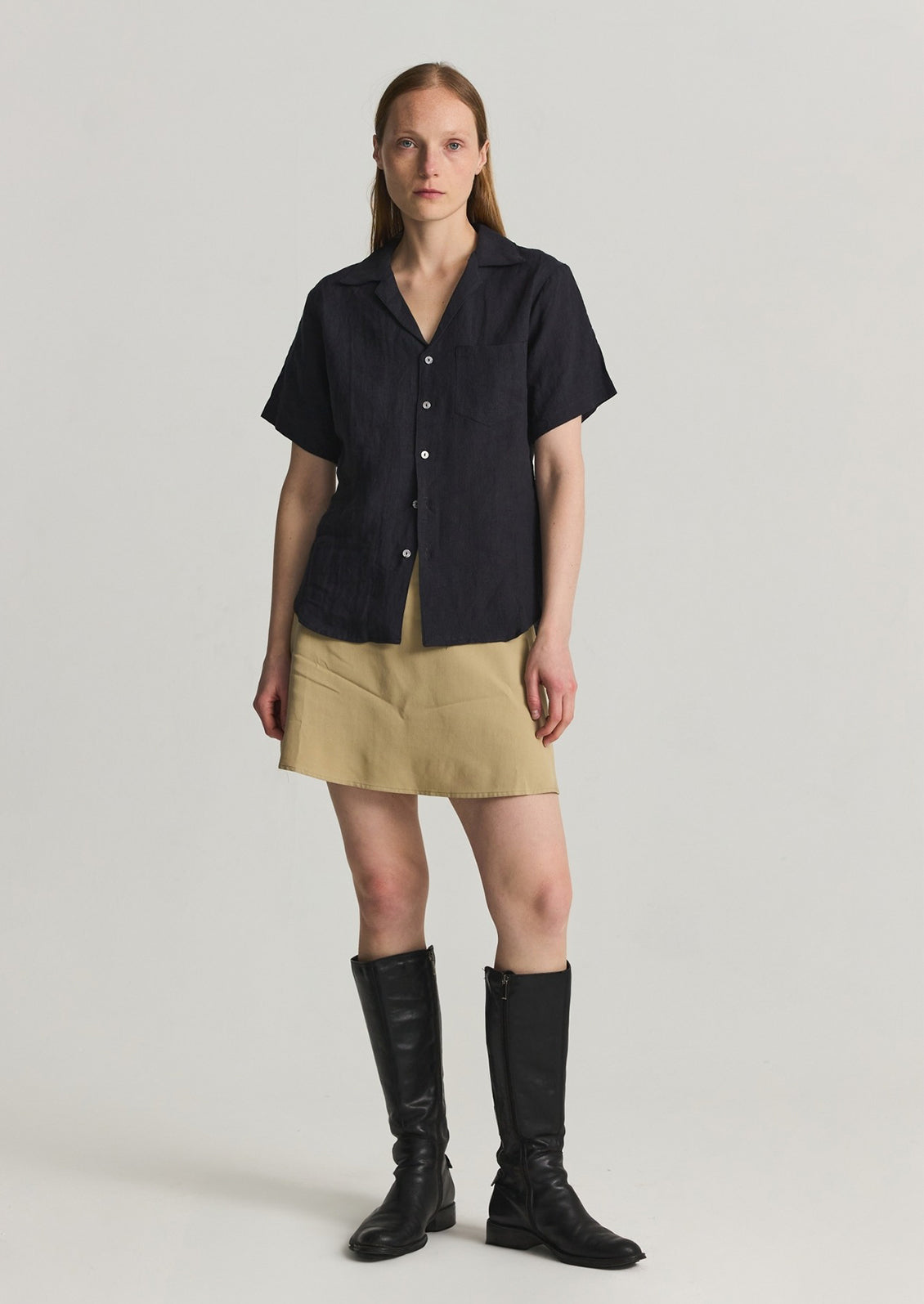 A woman wearing a black linen shortsleeved button-up, khaki skirt, and boots. 