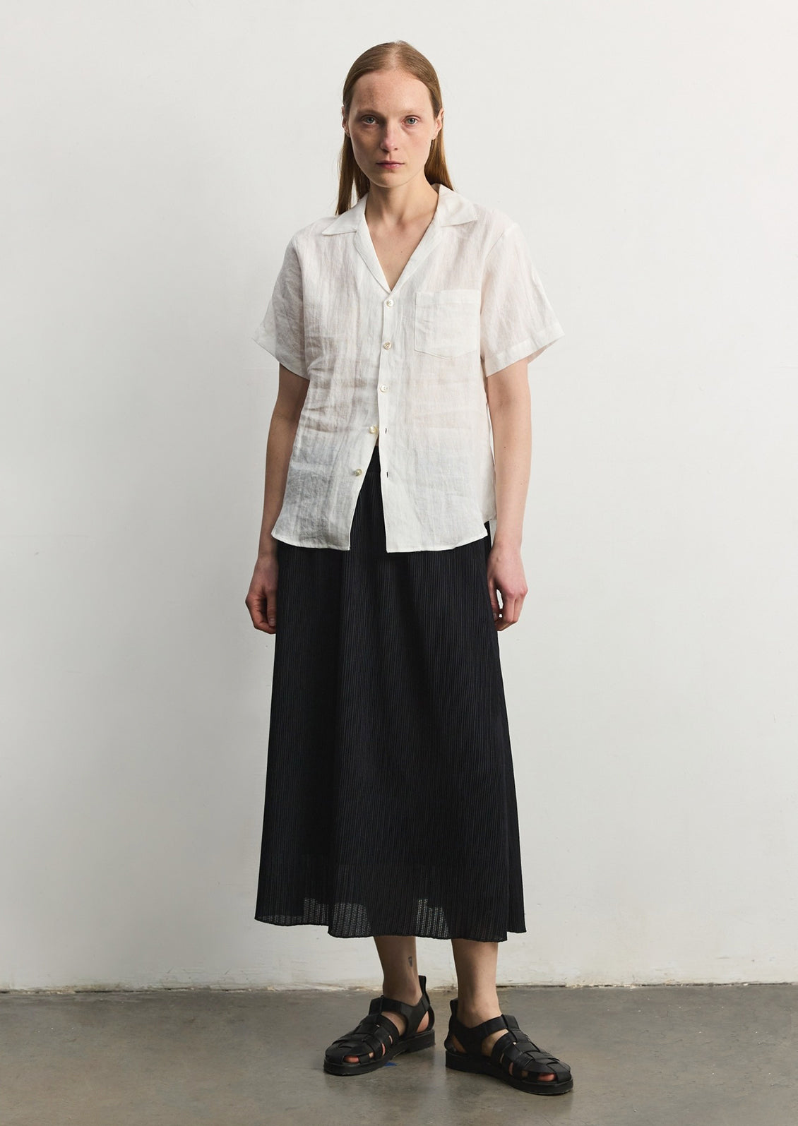 A woman wearing a white linen shortsleeved button-up, long black skirt, and sandals. 
