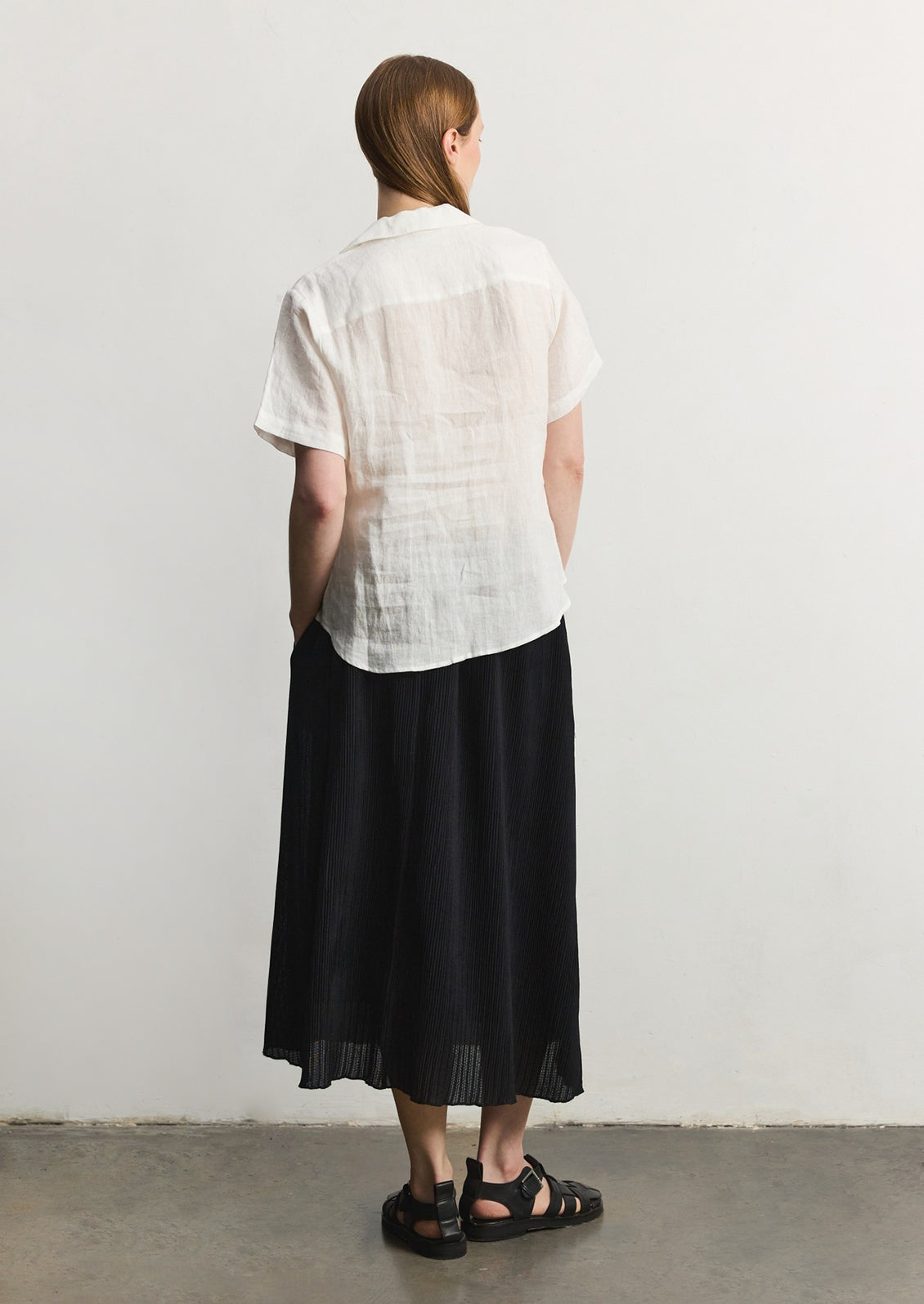 A woman wearing a white linen shortsleeved button-up shirt, dark skirt, and sandals, seen from behind. 