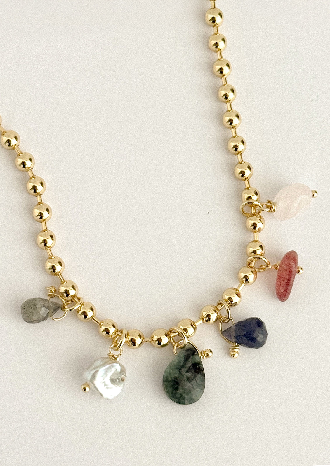 A gold beaded charm necklace featuring various raw, polished gemstone pendants in green, pink, blue, white, and gray, displayed on a light beige background.