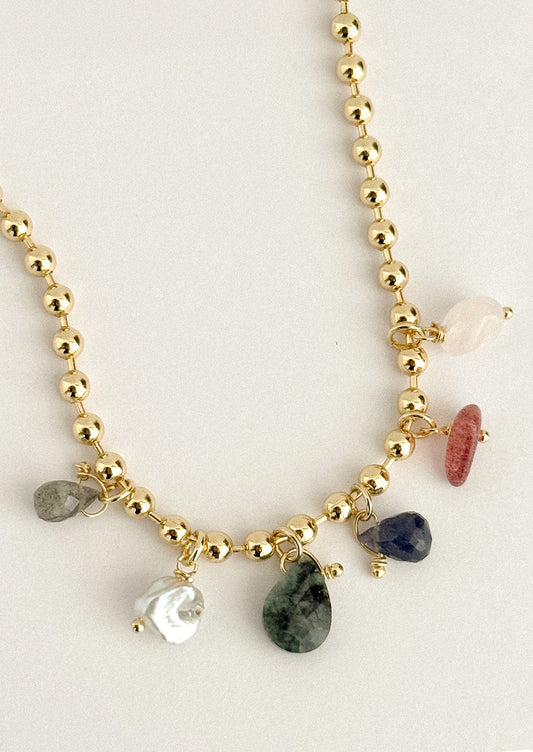A gold beaded charm necklace featuring various raw, polished gemstone pendants in green, pink, blue, white, and gray, displayed on a light beige background.