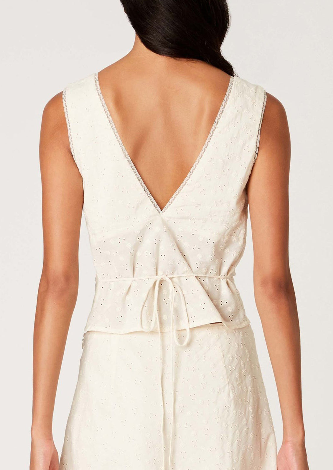 A woman wearing a sleeveless cotton eyelet lace top in cream, with tie back waist.