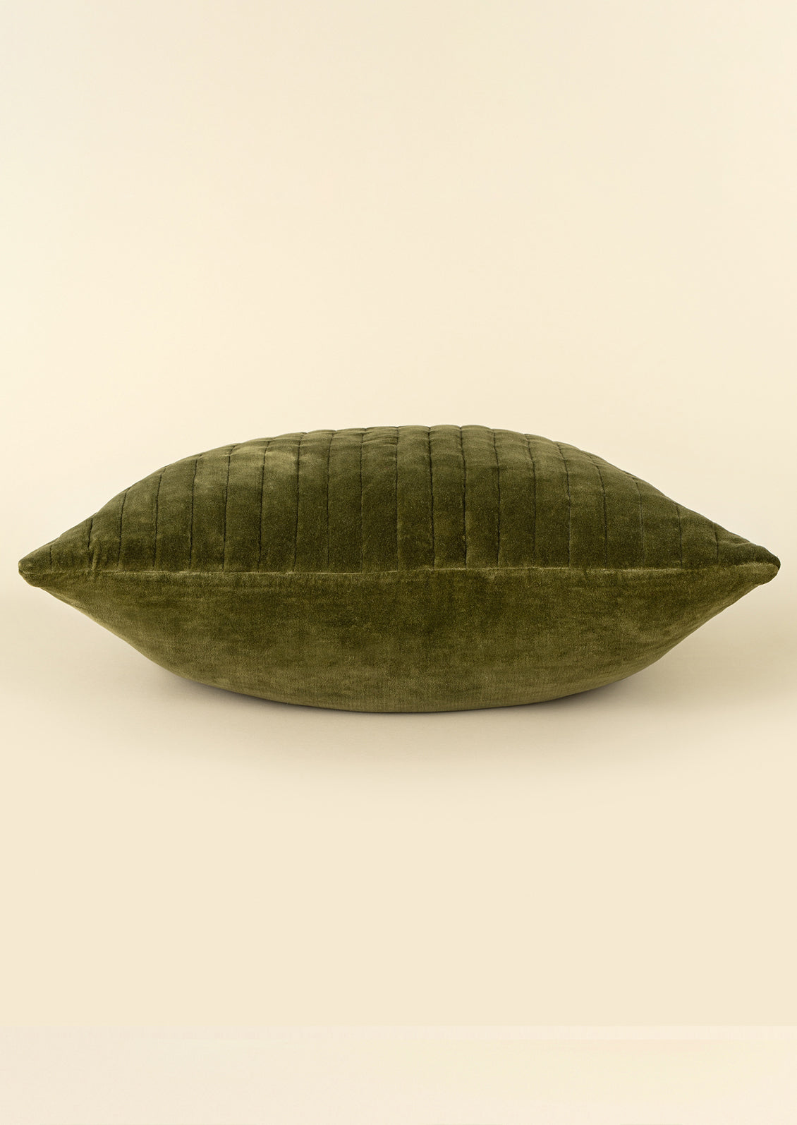 An olive green velvet pillow with vertical channel tufting on one side, shown against a solid cream background.