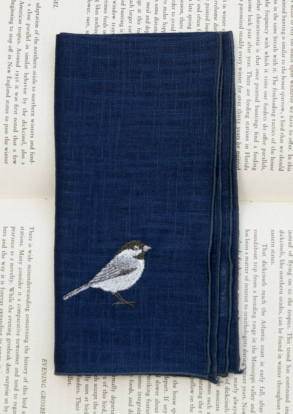 A navy blue linen napkin with tonal serged edges, and an embroidered bird at corner.
