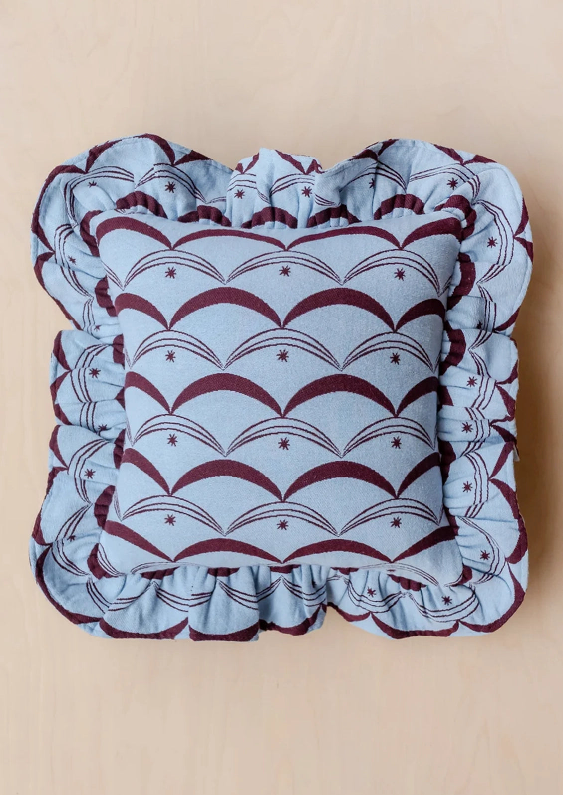 A square decorative throw pillow with a ruffled border, featuring a light blue and burgundy scallop pattern on a light wood background.