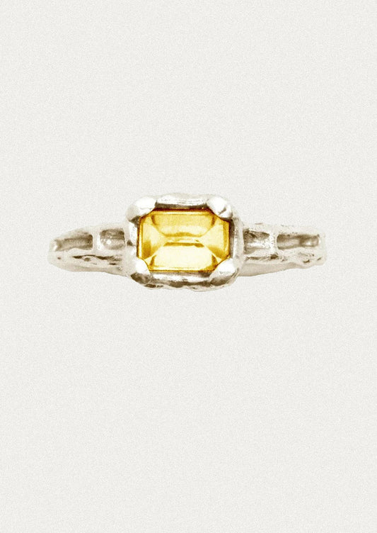 A silver ring featuring a rectangular yellow gemstone in a textured bezel setting, shown against a plain off-white background.