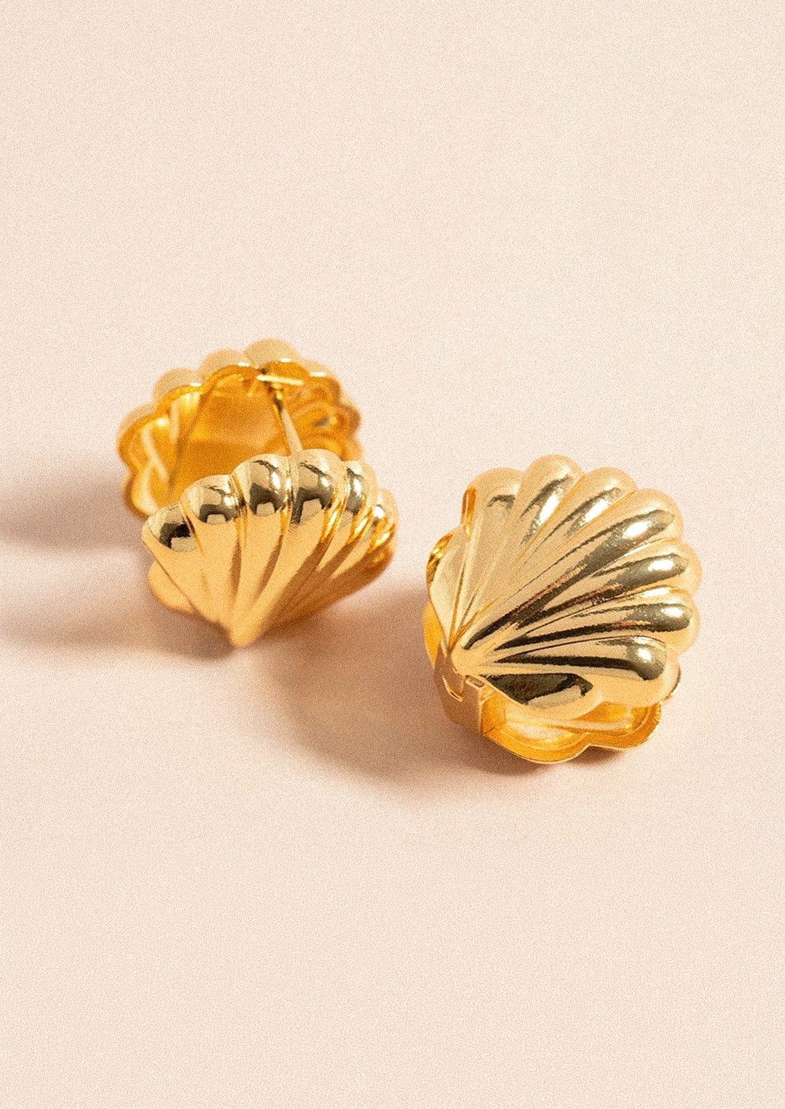 A pair of gold huggie earrings in the shape of a concha shell on a light pink background.