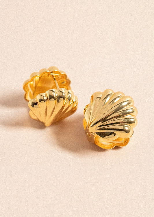 A pair of gold huggie earrings in the shape of a concha shell on a light pink background.