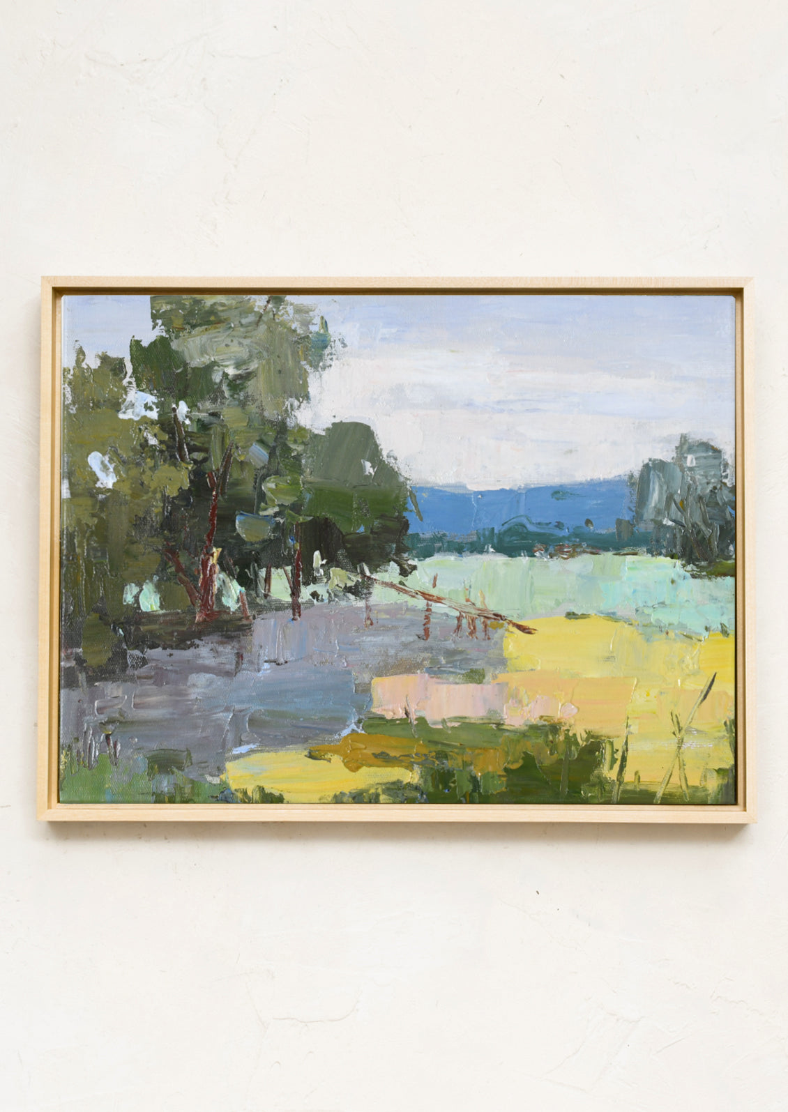 A framed landscape oil painting featuring green trees, a pale blue sky, and a bright yellow field, hung on a textured off-white wall.