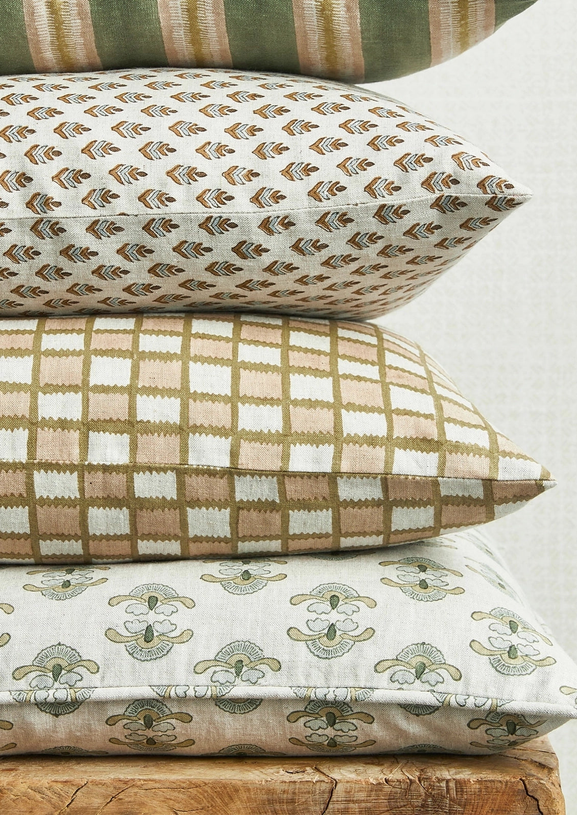 Three decorative throw pillows stacked on a rustic wooden surface against a light neutral background. The top pillow has a small repeating geometric pattern in brown and beige on an off-white base.