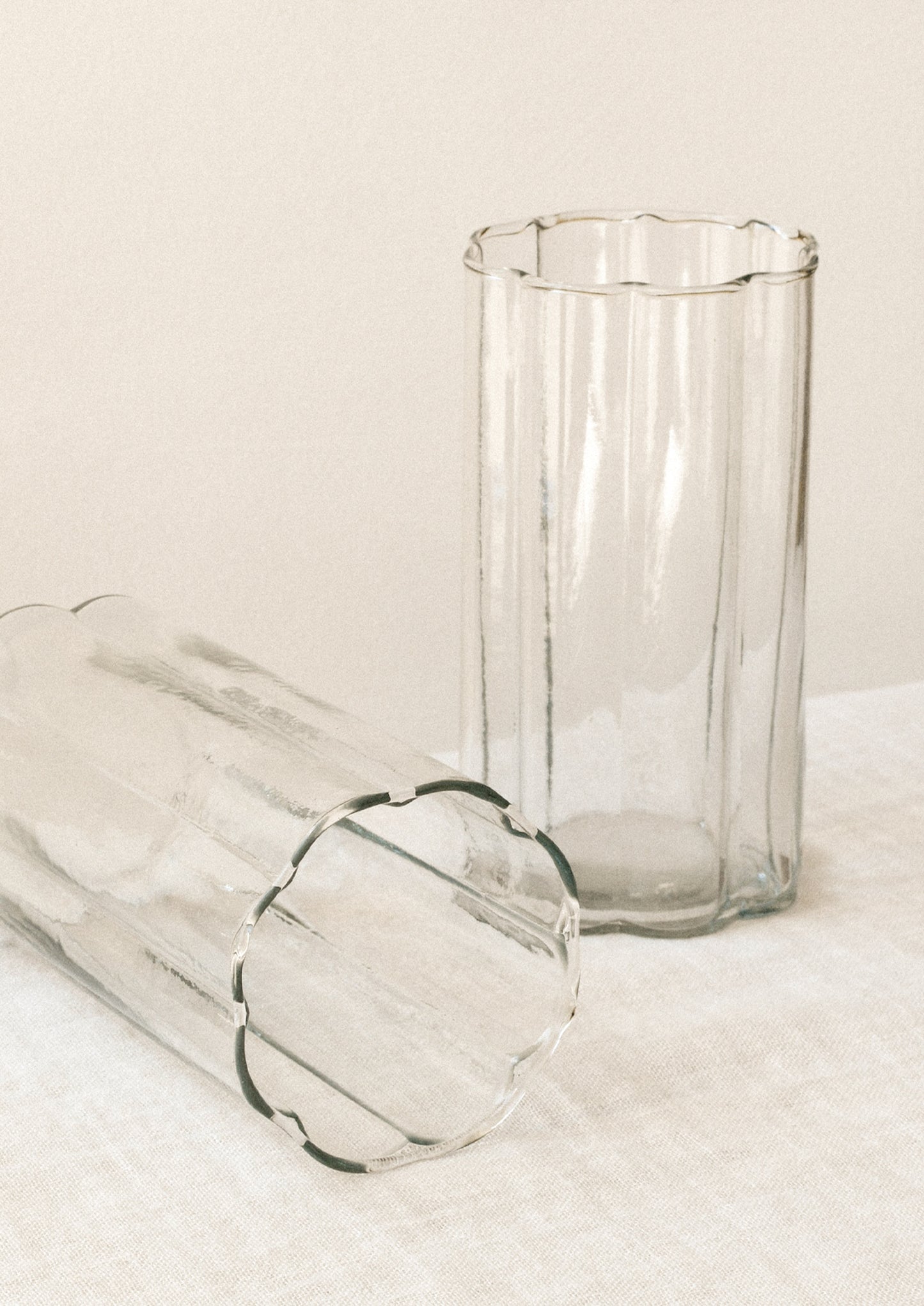 Two clear ribbed glass tumblers with scalloped edges, one standing upright and the other lying on its side, set against a warm off-white background.