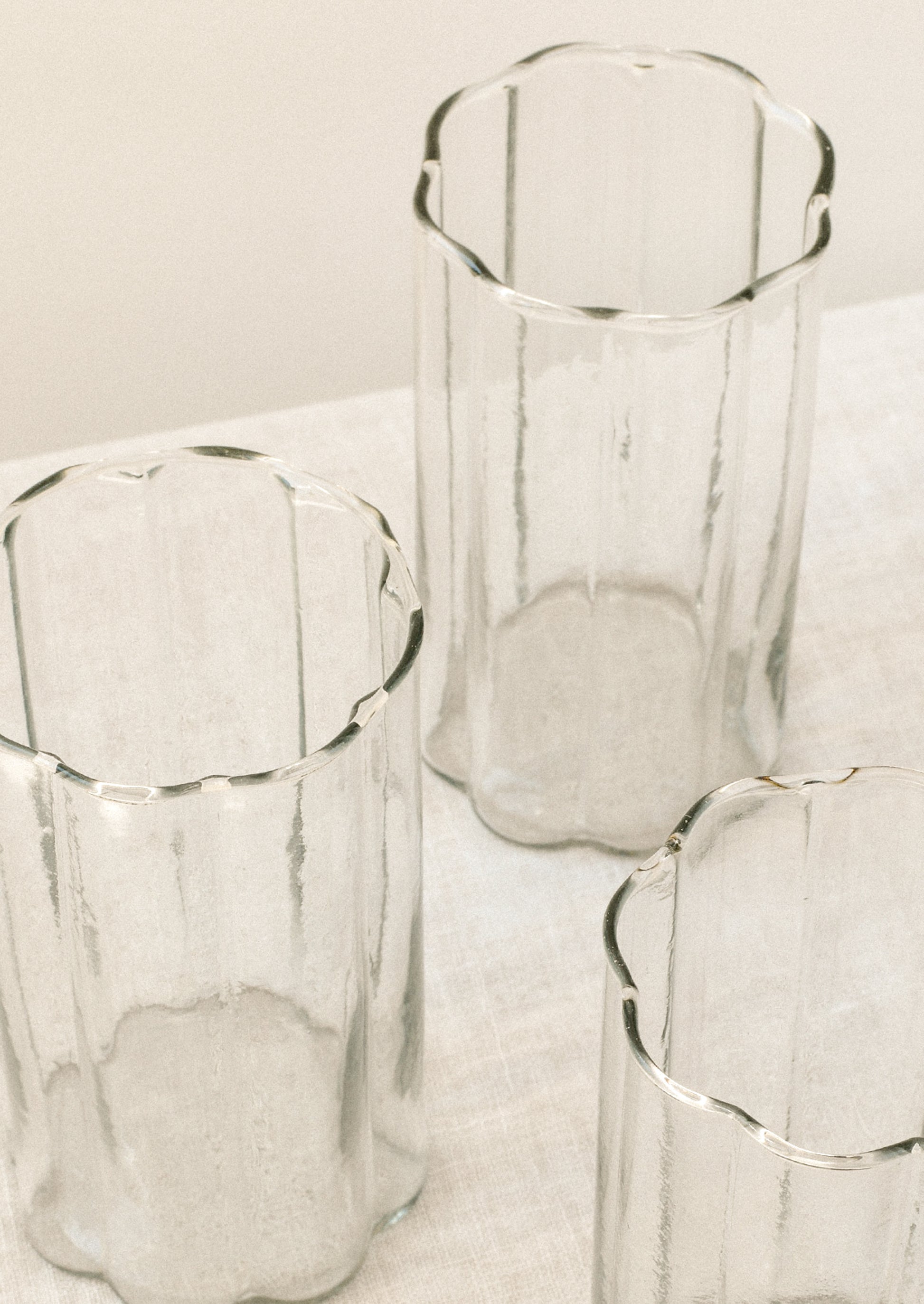 Three clear ribbed glasses with scalloped edges, arranged on a light fabric surface against a soft off-white background.