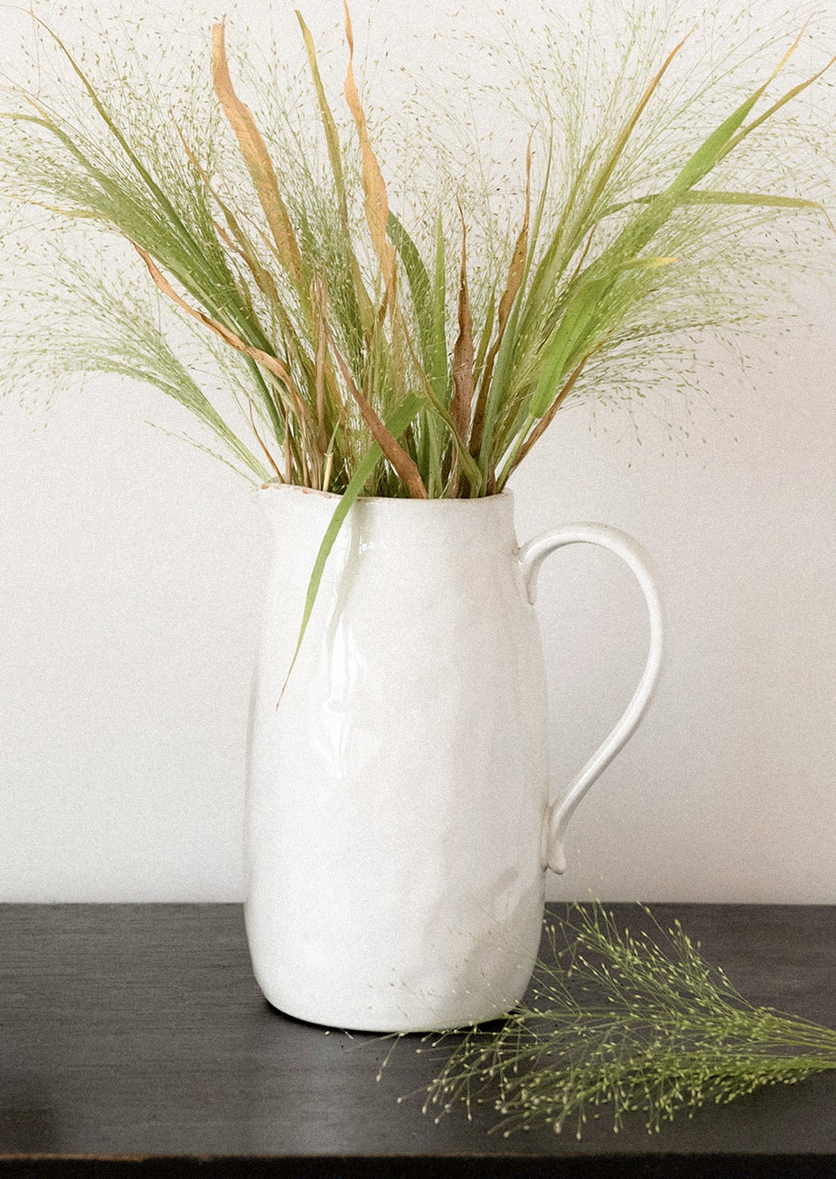 A white ceramic pitcher vase with a textured finish, holding dried green and tan grasses, set on a dark wooden surface against a light gray wall.