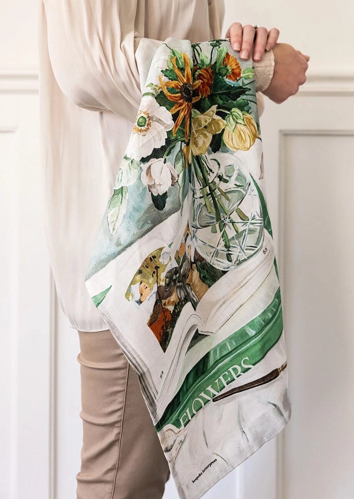 A linen tea towel features green stripes, white flowers, and sunflowers, with the word 'FLOWERS' printed on it.