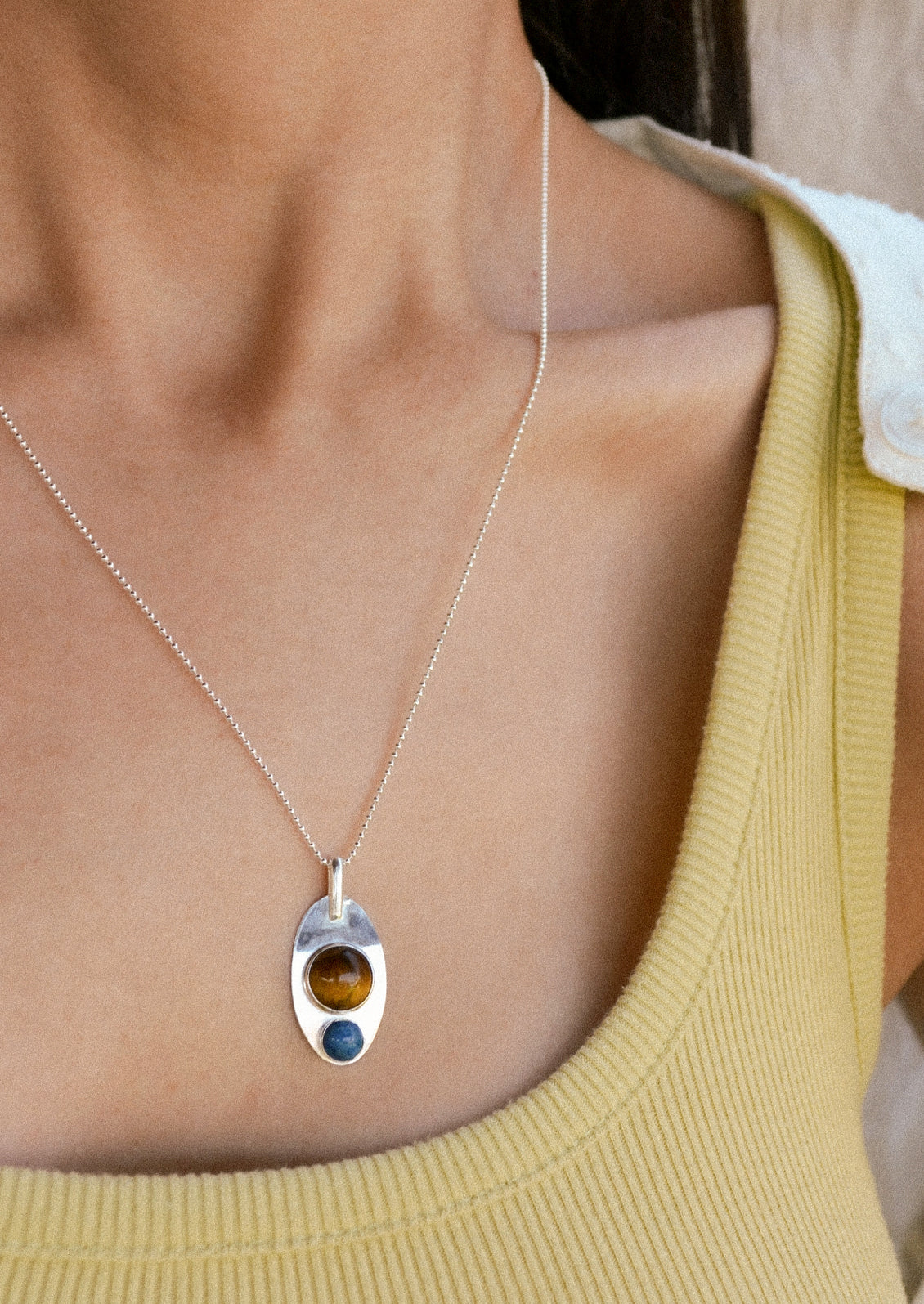 A woman wearing a silver necklace with pendant adorned with a brown gemstone and blue gemstone.