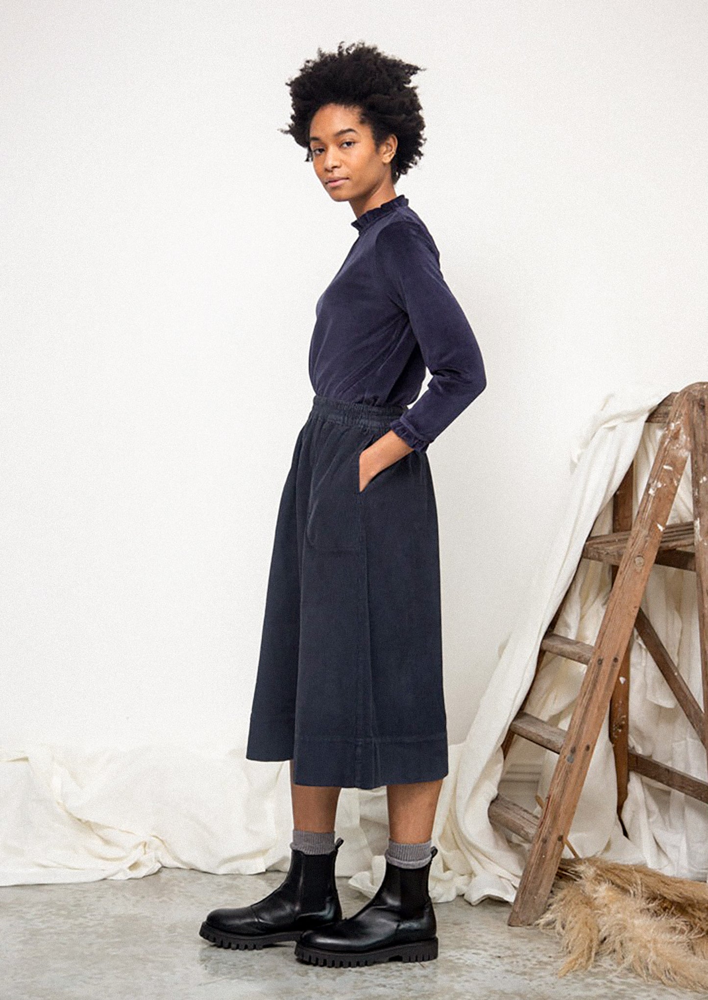 Woman wearing a navy blue dress standing in a minimalistic room with white curtains and wooden furniture.