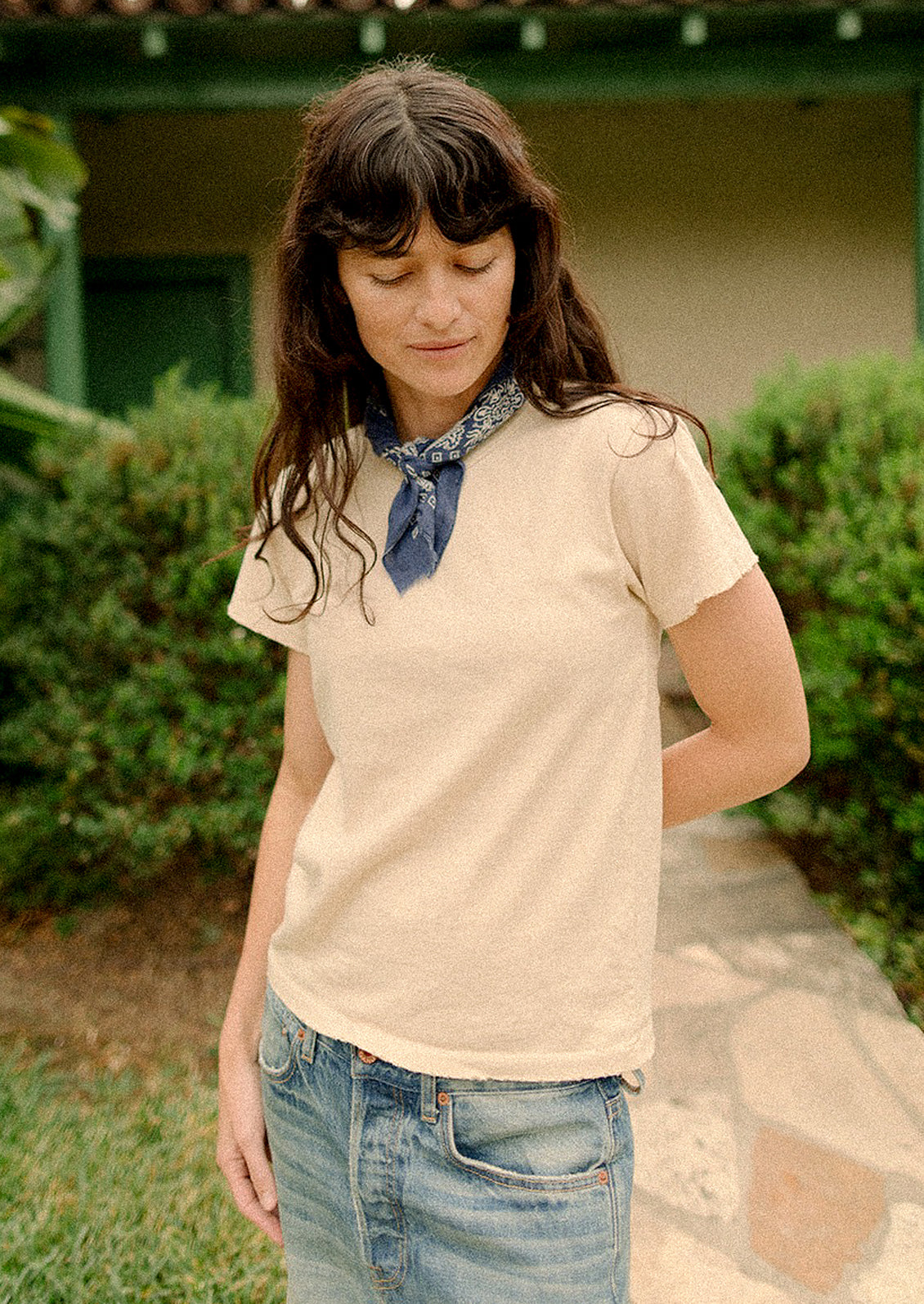 Woman wearing a beige shirt and blue jeans with a scarf, standing outdoors.