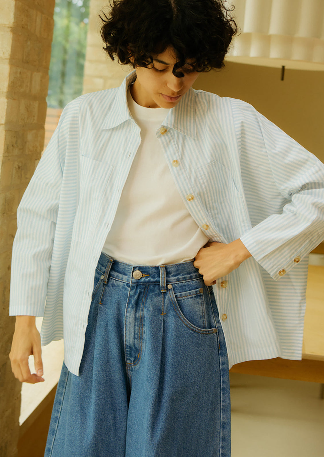 Person wearing a light blue striped shirt over a white sweater and blue jeans, standing indoors.